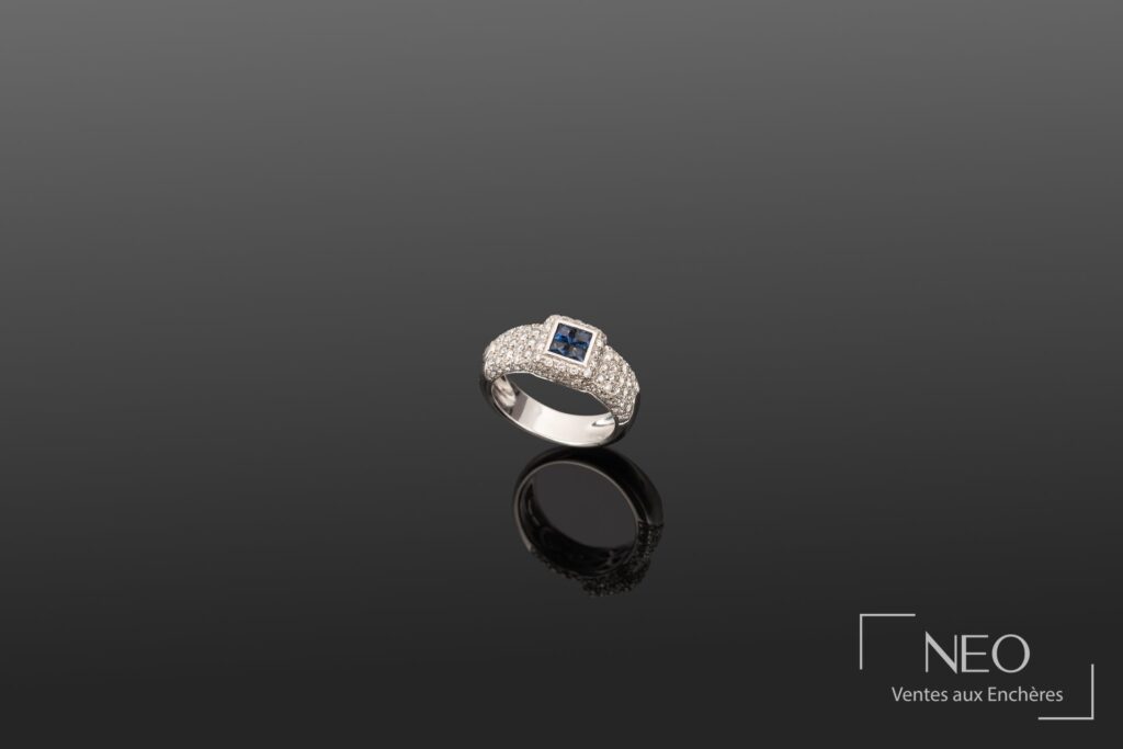 Bague en or gris sertie d'un motif carré avec saphirs et diamants sur fond sombre.