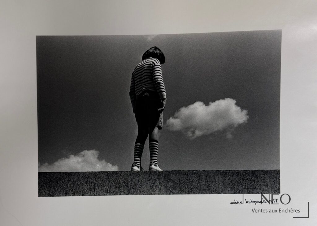 Photographie en noir et blanc d'un enfant debout sur un muret sous un ciel nuageux