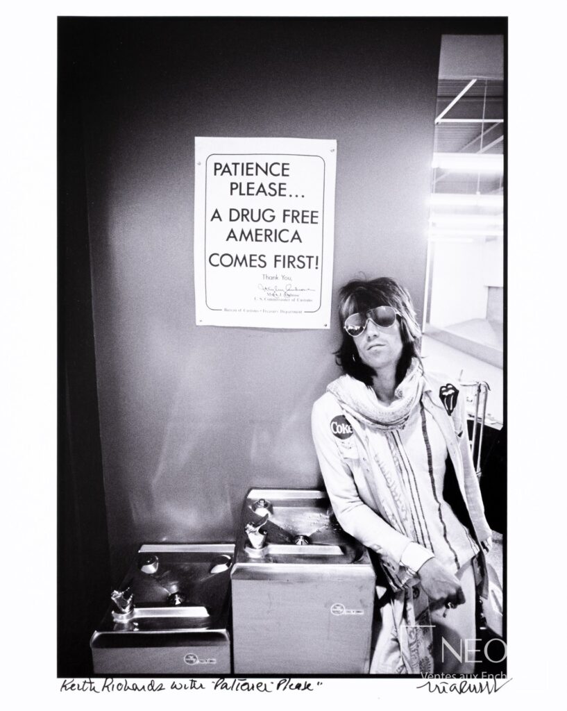 Photographie noir et blanc de Keith Richards devant une affiche « Patience Please »