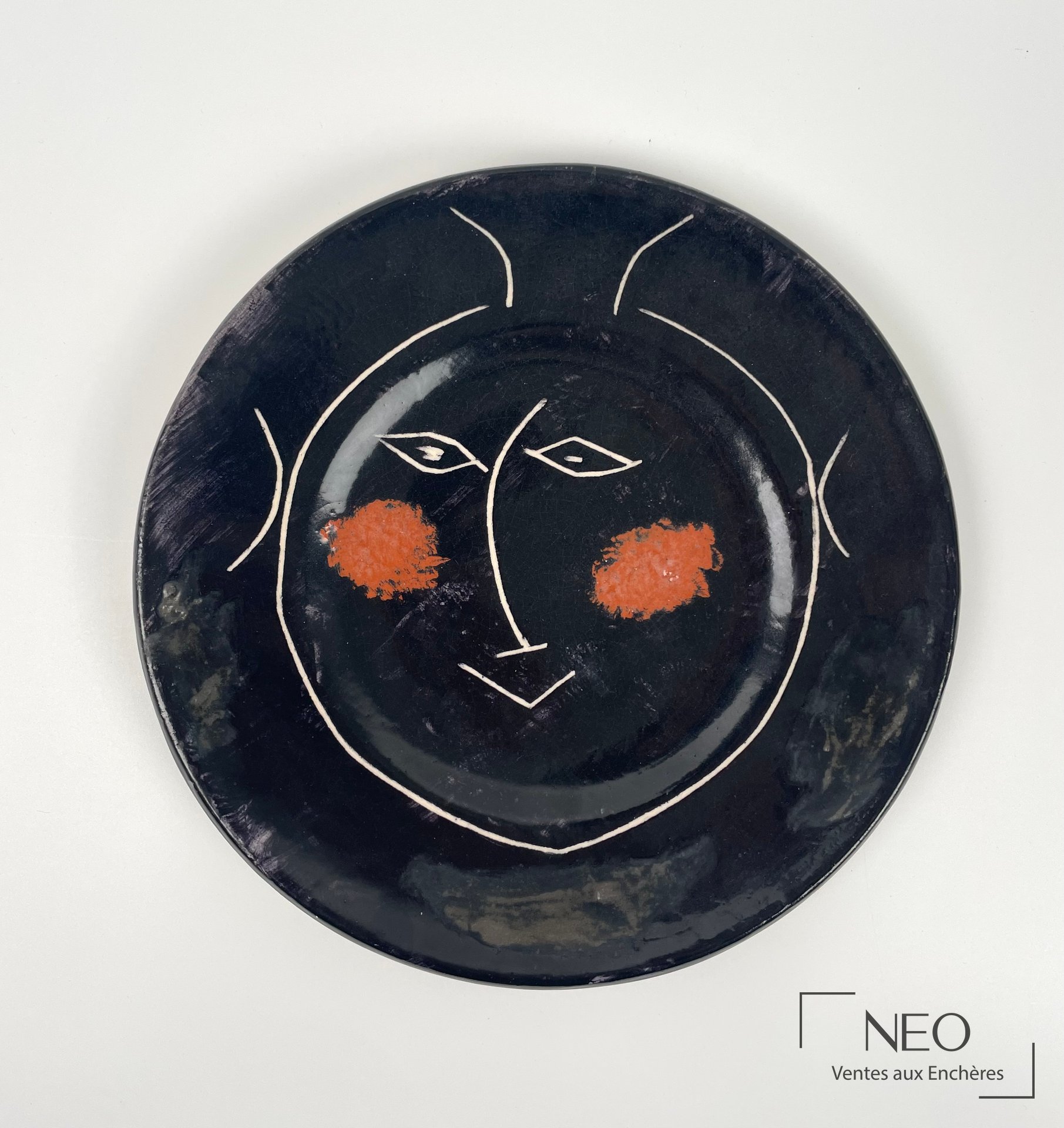 Assiette noire décorée d'un visage stylisé avec deux taches orange et des traits blancs
