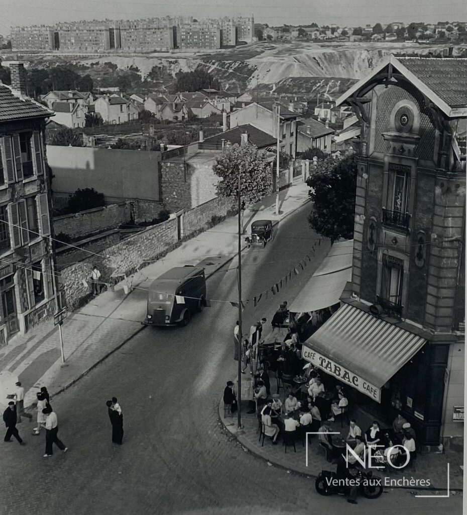 Photographie en noir et blanc d'une rue en hauteur avec café d'angle et passants.