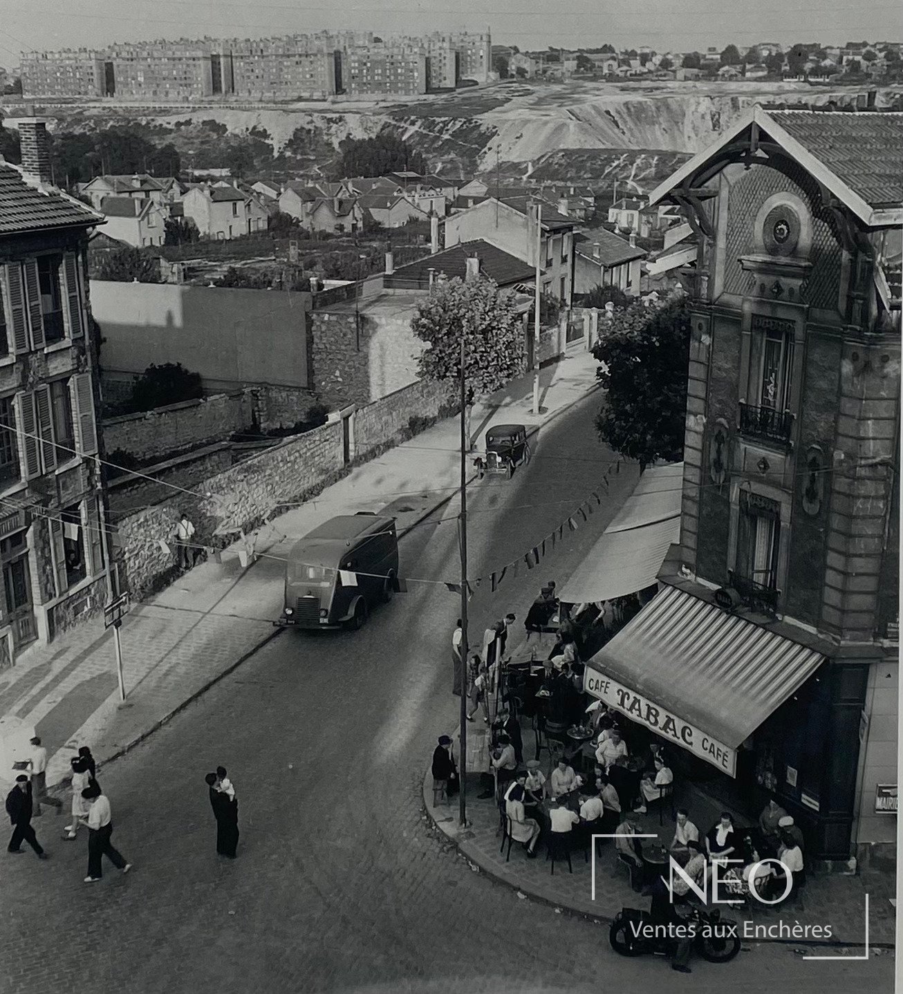 Photographie en noir et blanc d'une rue en hauteur avec café d'angle et passants.